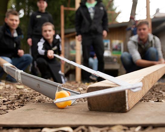 Eine Gruppe junger Menschen machen ein Experiment mit einem Holzbalken, einer Regenrinne, einem Zollstock und einer Quietsche Ente