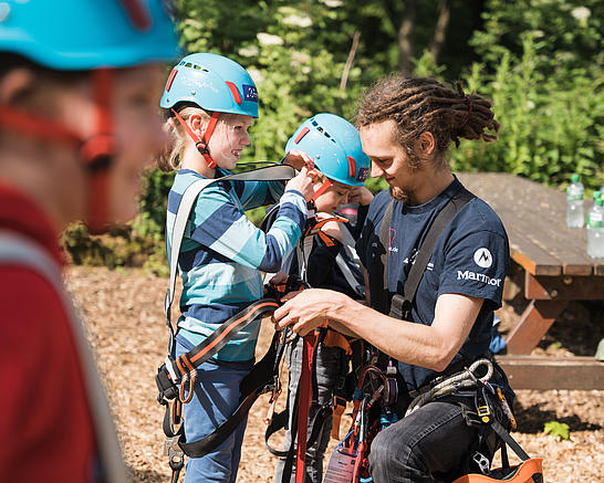 Ein Mitarbeiter des Kletterparks hilft Kindern beim Anziehen der Klettergurte
