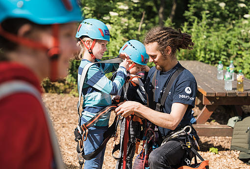 Ein Mitarbeiter des Kletterparks hilft Kindern beim Anziehen der Klettergurte