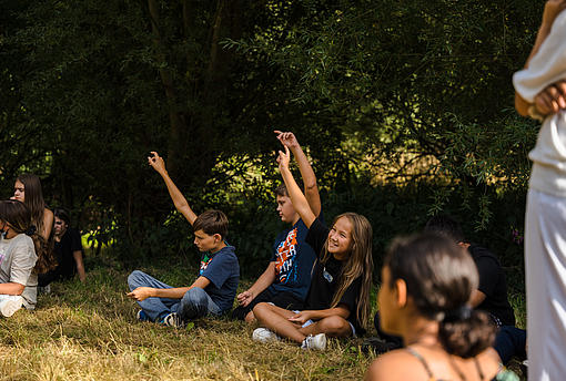 Kinder melden sich während einer Feedbackrunde auf der Wiese sitzend.