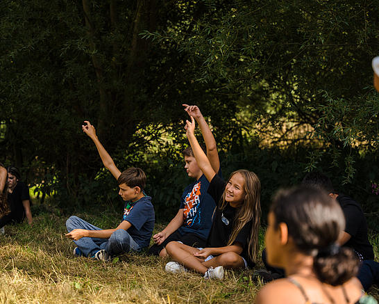 Kinder melden sich während einer Feedbackrunde auf der Wiese sitzend.