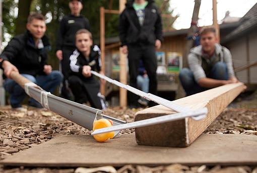 Eine Gruppe junger Menschen machen ein Experiment mit einem Holzbalken, einer Regenrinne, einem Zollstock und einer Quietsche Ente